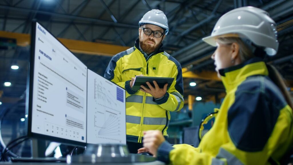 Two assemblers with helmets, one sitting in front of computer screen, the other one standing and making notes